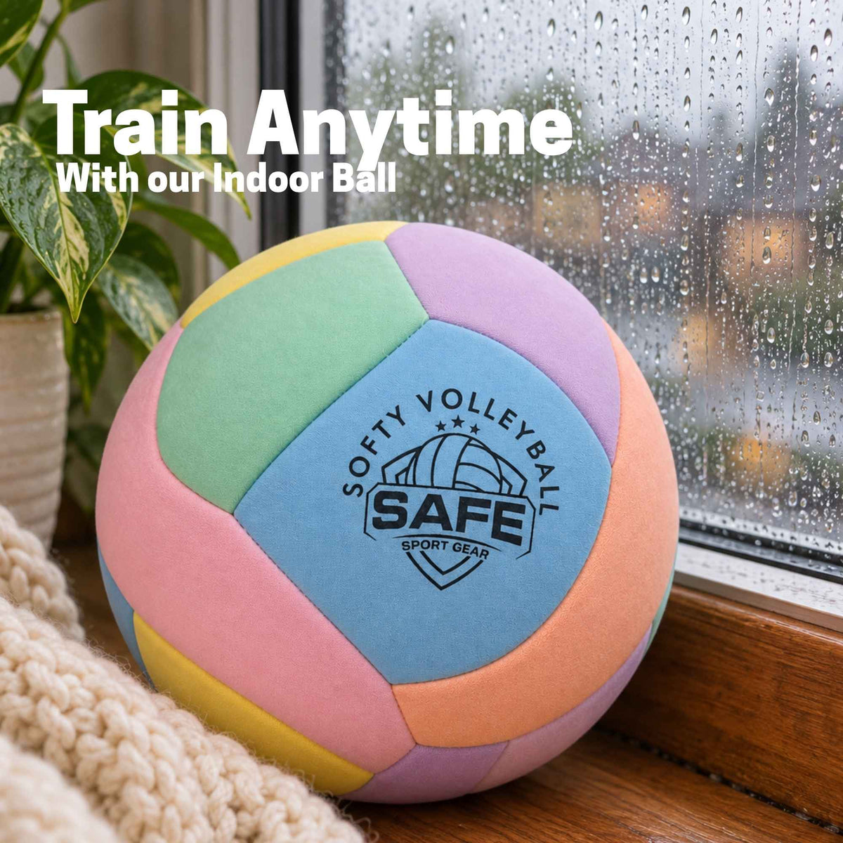 A pastel-colored indoor volleyball with the text 'SOFTY VOLLEYBALL' printed on it, resting on a windowsill with raindrops on the glass and a plant in the background. White text overlay reads 'Train Anytime With our Indoor Ball'.