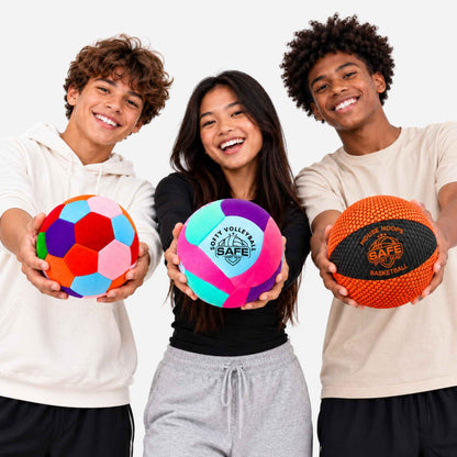 Three smiling young athletes hold colorful houseball, volleyballs and basketballs against a clean white studio background. The center child holds a bright pink, purple, and teal ball with 'SAFE Sport Gear' logo printed on it.