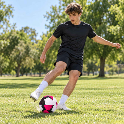 A young man in a black athletic t-shirt and black shorts kicks a pink, white, and black soccer ball on a green grass field with trees in the background.