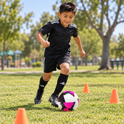 A young boy wearing a black soccer jersey, shorts, and socks dribbles a purple, white, and black soccer ball on a green grass field, with orange cones and trees in the background.