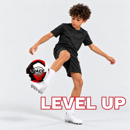 A young boy with curly brown hair is shown kicking a red soccer ball in a studio setting. He is wearing a black t-shirt, black shorts, white socks, and white soccer cleats.