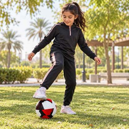 A young girl in a black athletic tracksuit with pink accents is playing with a red pace raptor soccer ball on a sunny grassy park lawn.