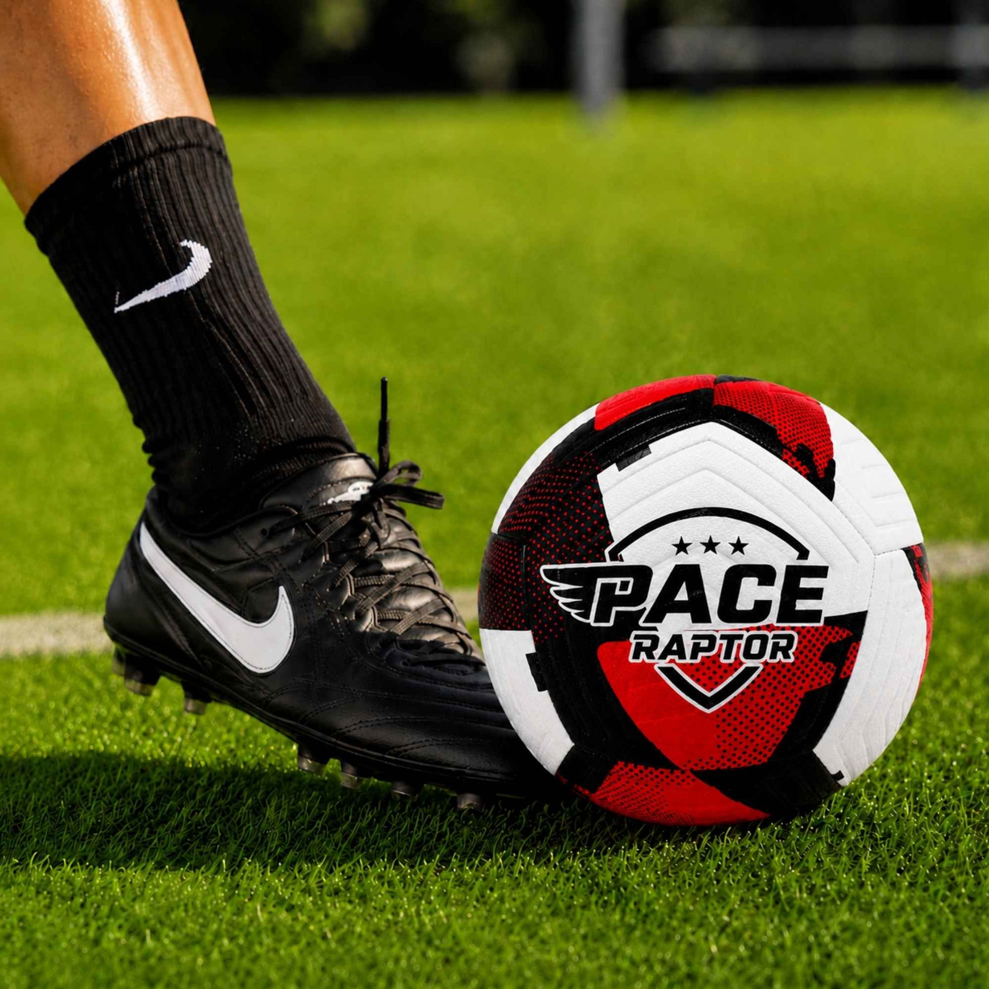 Pace Raptor Size 4 Soccer Ball - Red with 'Pace Raptor' logo on a grass field, close-up of foot wearing black soccer cleats