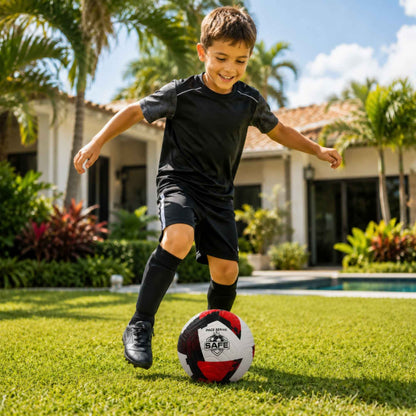 Child playing with a Pace Raptor Size 4 Soccer Ball - Red  on a grassy lawn with a house and palm trees in the background