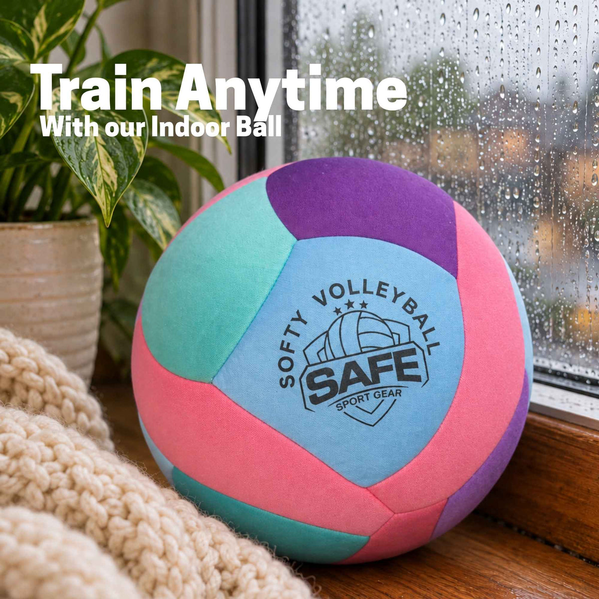 A pastel-colored indoor ball volleyball with pink, teal, light blue, and purple panels sits on a windowsill with a knit blanket nearby. The ball has a 'SOFTY VOLLEYBALL SAFE SPORT GEAR' logo printed on it.