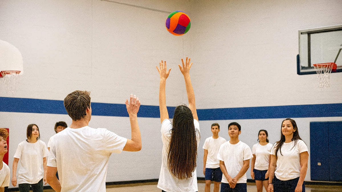 A group of people in a gymnasium, with a woman in the center reaching up to hit a colorful ball on a basketball hoop, wearing matching white t-shirts.