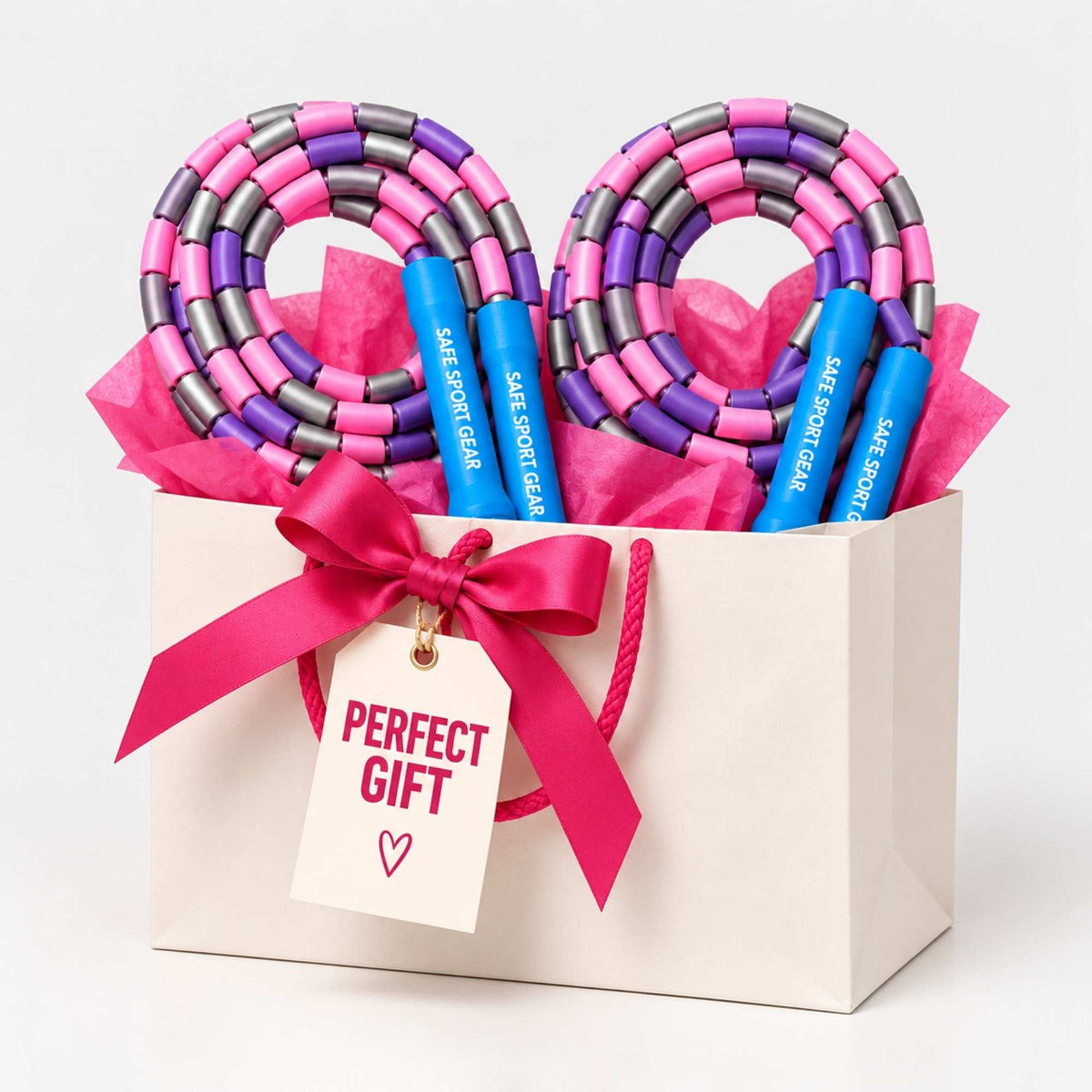 A gift set in a white paper bag with a bright pink ribbon and tag reading 'PERFECT GIFT'. Inside the bag are two pink and purple jump ropes with blue handles labeled 'SAFE SPORT GEAR'.