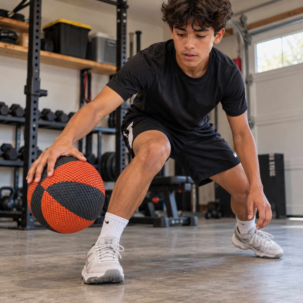 A man in a black athletic t-shirt and black shorts crouches in a garage gym holding an orange and black basketball, wearing white Nike sneakers and white Nike socks.