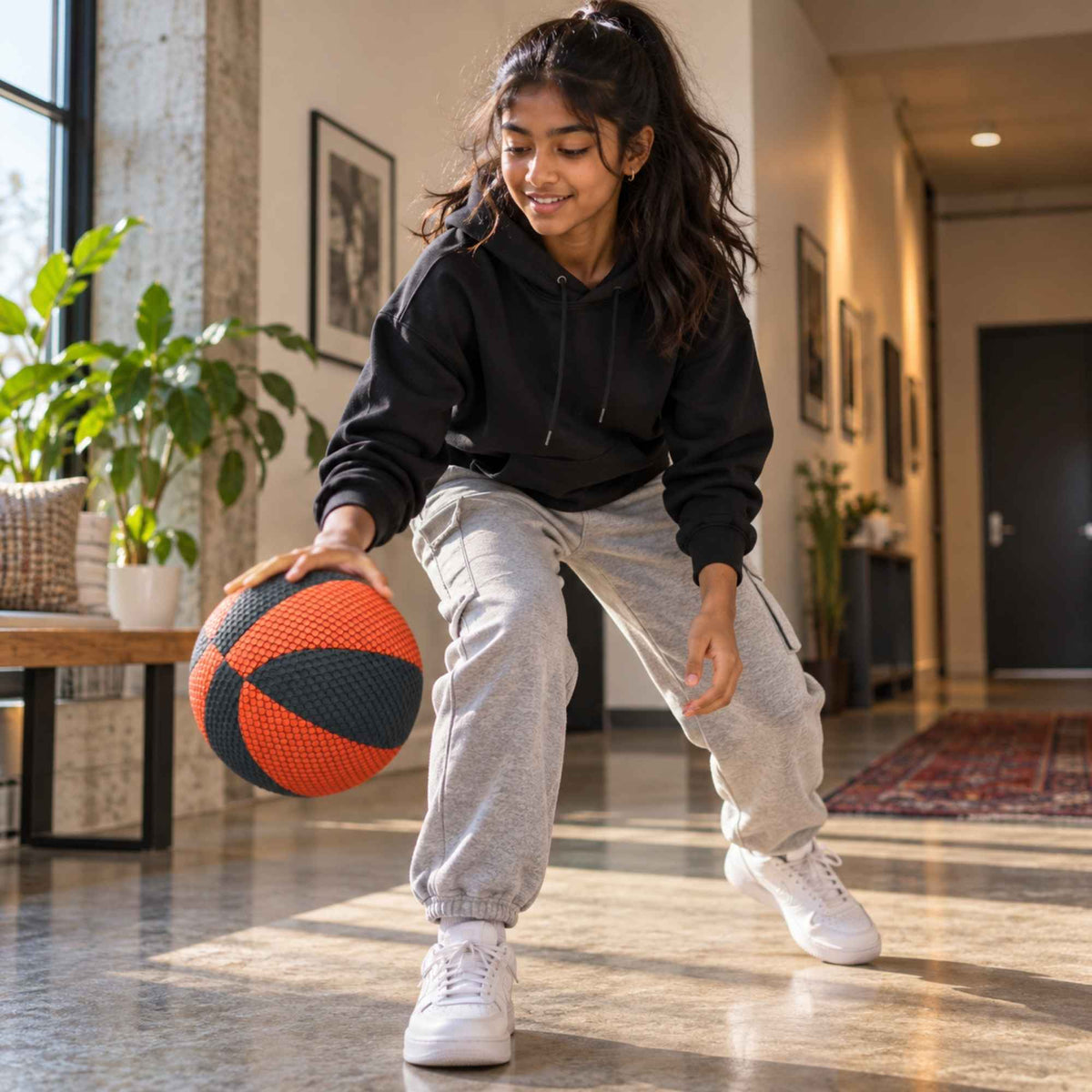 A young girl wearing a black hoodie, light gray sweatpants, and white sneakers is crouching indoors while holding a househoops basketball in a brightly lit hallway.