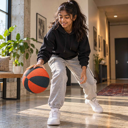 A young girl wearing a black hoodie, light gray sweatpants, and white sneakers is crouching indoors while holding a househoops basketball in a brightly lit hallway.